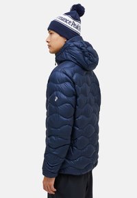 Navy blue puffy jacket with quilted wave pattern, hood, and logo on the sleeve. Worn with a matching navy and white pom-pom beanie.