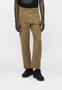 Pantaloni din catifea maronii cu un design drept, având un talie cu nasturi și o textură de riburi vizibilă. Purtați cu adidași negri.