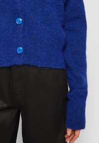 Blå ullkofta med två ljusblå knappar, en kort design, ribbade ärmslut och en mjuk texturerad yta. Matchad med svarta byxor.