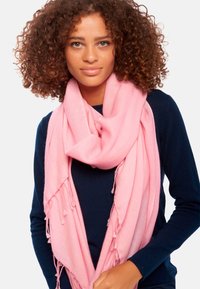 Écharpe rose en tissu doux, avec des accents de franges. Portée lâchement autour du cou, associée à un haut bleu foncé.