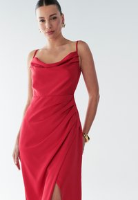 Červené saténové šaty s širokým vodopádovým výstrihom, tenkými ramienkami a postranným záhybom. Majú vysoký rozparok na stehne a hladkú textúru. Zlaté doplnky.