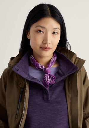 Braune Jacke über einem violetten, texturierten Hemd mit Knopfleiste, kombiniert mit einem gemusterten violett-weißen Schal, der am Hals gebunden ist.