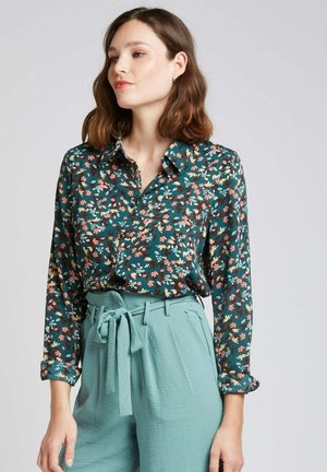 Chemise à motifs floraux à boutons en vert foncé avec des motifs roses et bleus, associée à un pantalon bleu clair avec une taille nouée et un tissu texturé.