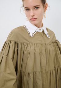 Abito camicia oversize verde oliva con colletto bianco a scallop, design plissettato e chiusura con bottoni, realizzato in un morbido tessuto testurizzato.