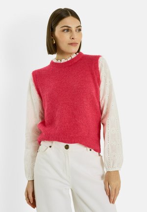 Femme portant des boucles d'oreilles créoles en or, un gilet tricoté sans manches rose vif par-dessus un chemisier blanc à manches longues avec des détails en broderie anglaise, et un pantalon blanc.