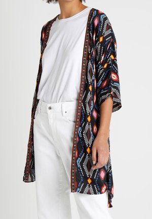 Henkilö, joka pukeutunut valkoiseen t-paitaan ja housuihin sekä mustaan kimono-tyylisten jakkuun, jossa on värikkäitä geometrisia kuvioita.