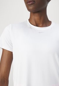 Camiseta deportiva blanca de manga corta, con cuello redondo y un sutil logo de Nike en gris en el pecho. Tejido suave y ligero.