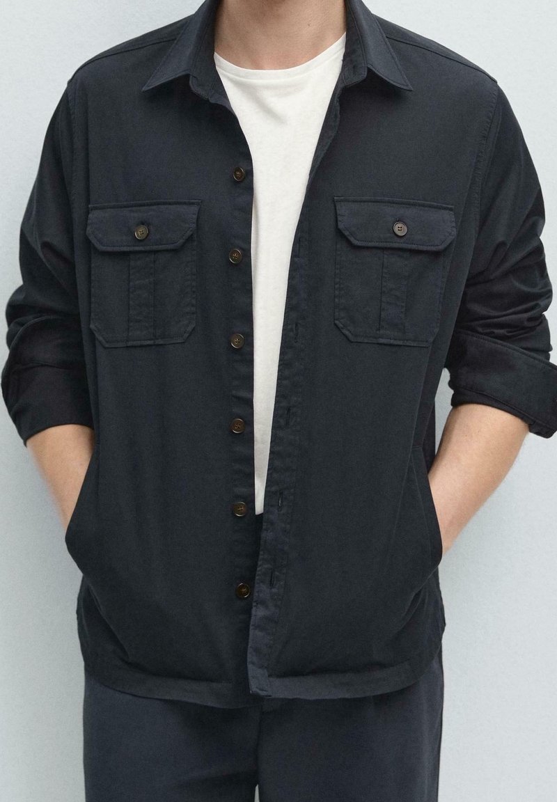 Personne portant une veste noire boutonnée avec deux poches avant sur une chemise blanche, mains dans les poches de la veste et manches retroussées.