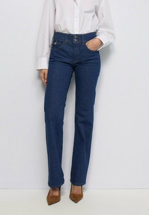 Mujer con vaqueros azul oscuro de tiro alto y pierna recta, camisa blanca abotonada y tacones marrones de punta afilada, de pie contra una pared lisa.