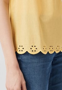 Geel stoffen top met een geschulpte zoom en bloemenvormige uitsnijdingen. Gepaard met blauwe jeans, wat een casual ontwerp toont.