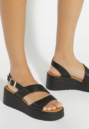 Pies con sandalias de plataforma negras con dos tiras anchas y una correa en el tobillo con una hebilla dorada, sobre un fondo blanco.
