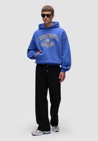 Niebieska bluza z kapturem z napisem "WRSTBHVR STUDIO" w białym i czarnym kolorze, w połączeniu z czarnymi spodniami. Szare i białe sneakersy dopełniają stylizację.