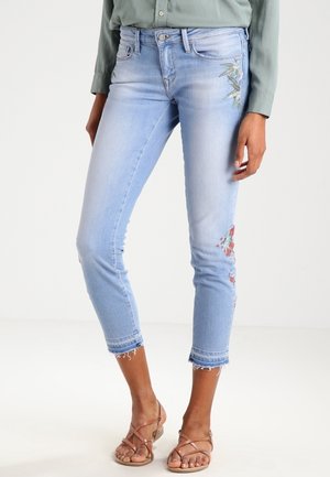 Vaqueros crop de color azul claro de corte ajustado con bordado floral cerca del bolsillo y la rodilla, combinados con sandalias beige de tiras y camisa verde de manga larga.