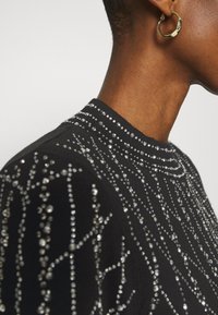 Pull noir avec un col montant, orné d'ornements argentés disposés en motif dispersé, créant une surface texturée.