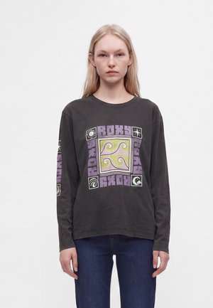Jeune femme aux cheveux blonds et raides porte un t-shirt noir à manches longues avec un motif violet et jaune et un jean bleu, debout sur fond blanc.