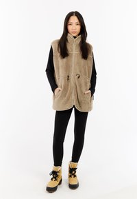 Gilet in pile beige con colletto alto, zip frontale e due tasche, indossato sopra una maglietta nera a maniche lunghe e abbinato a leggings neri e stivaletti gialli.