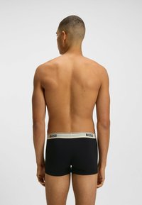 Man som står vänd bort, iklädd svarta boxerkalsonger med en vit midja märkt "BOSS" mot en enkel ljus bakgrund.