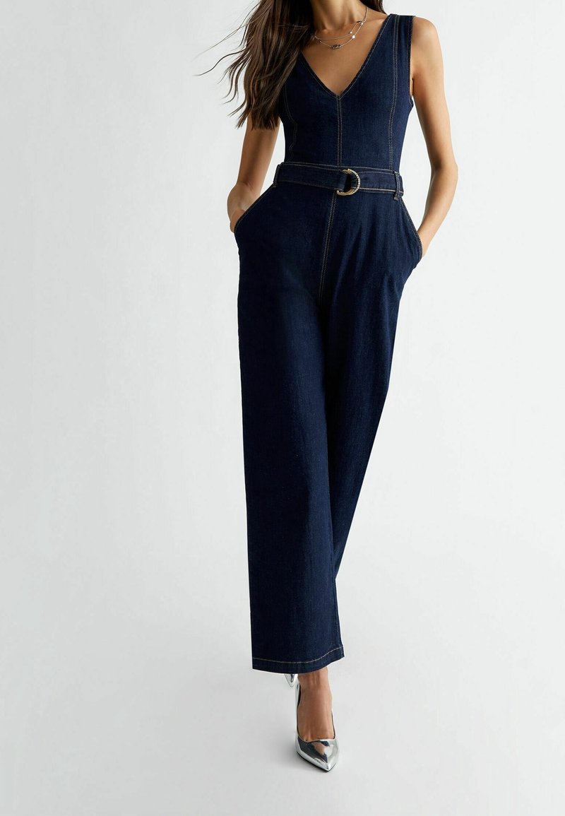 Femme portant une combinaison en denim foncé sans manches avec col en V, taille ceinturée, jambes larges, mains dans les poches et talons aiguilles argentés pointus.