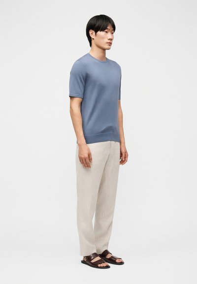 Άνδρας που στέκεται, φοράει μπλε πουκάμισο με κοντά μανίκια, ανοιχτό μπεζ παντελόνι και σκούρα καφέ σανδάλια, μπροστά σε απλό ανοιχτό φόντο.