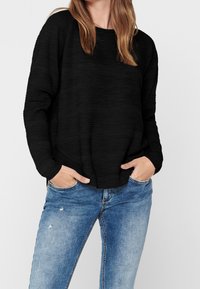 Pullover nero con texture, dal taglio morbido, colletto rotondo e maniche lunghe, abbinato a jeans in denim blu strappati.