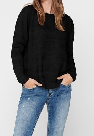 Pull noir texturé à coupe ample, col rond et manches longues, associé à un jean en denim bleu déchiré.