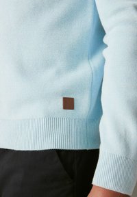 Šviesiai mėlynas megztinis su raštinėmis rankovėmis ir apatine dalimi, su mažu rudu odiniu ženkleliu apatiniame kairiajame kampe. Faktūruota medžiagos paviršius.