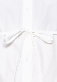 Vitt skjortan med knytband i midjan, tillverkad av ett lätt och slätt material med klara knappar.