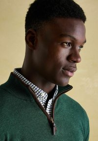 Zaļš džemperis ar brūnu rāvējslēdzēja apkakli virs zila un balta rūtainā krekla. Tuvplāns uz materiāla tekstūru un detaļām.