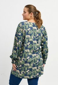 Langärmlige Tunika in einem geometrischen Muster aus Grün und Marineblau, aus weichem Stoff mit lockerer Passform und gerafften Bündchen, kombiniert mit blauen Jeans.