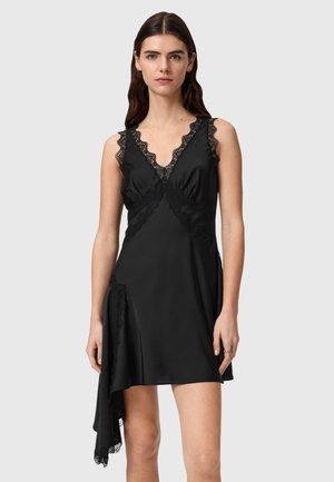 Vrouw draagt een mouwloze zwarte satijnen jurk met kanten rand en een asymmetrische zoom, staand tegen een effen lichtgrijze achtergrond.