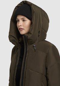 Žena v čiernej pletenej čiapke a tmavozelenej pérovke s veľkou kapucňou, čiastočne zapnutou, na jednofarebnom pozadí.