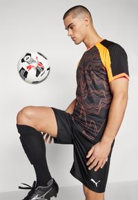 Puma sort og orange sportsdragt med bølget rødt mønster, kombineret med sorte shorts og knæhøje sokker, der viser en sort og hvid fodbold.