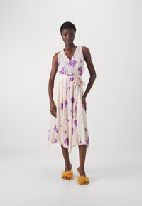Abito senza maniche di colore crema con motivo floreale, scollatura a V e vita con cintura; presenta fiori viola e una gonna fluida. Indossato con sandali arancioni pelosi.