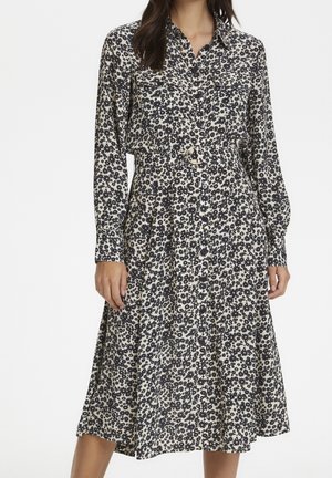Femme portant une robe fleurie noire et blanche à manches longues, boutonnée, avec une taille cintrée et une ceinture à boucle ronde dorée.