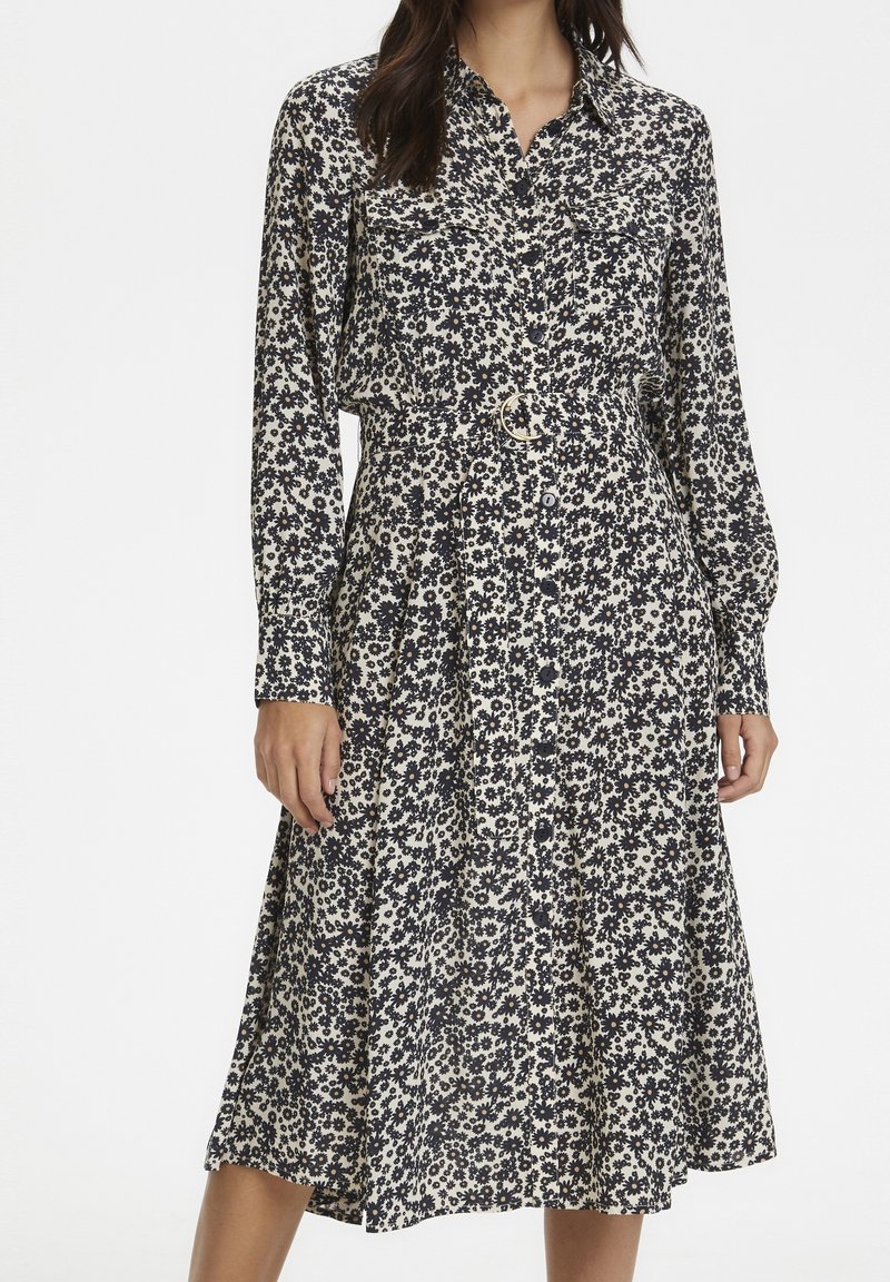 Femme portant une robe fleurie noire et blanche à manches longues, boutonnée, avec une taille cintrée et une ceinture à boucle ronde dorée.