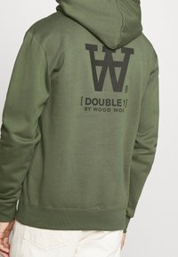 Sudadera verde hecha de una suave mezcla de algodón. Presenta un gran logotipo gráfico negro en la parte posterior. Incluye capucha con cordón y puños acanalados.