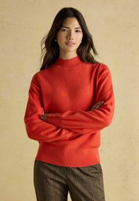 Roter Strickpullover mit hochgeschlossenem Kragen und mit Knöpfen verzierten Bündchen, kombiniert mit braunen karierten Hosen. Weicher Stoff, gerippter Saum und Bündchen.