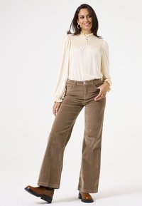 Crème blouse met lange mouwen, voorzien van smockdetails en een knoopsluiting, gecombineerd met high-waisted bruine flared corduroypants en bruine enkellaarzen.