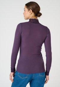 Maglietta a maniche lunghe di colore viola con una texture a coste; presenta un colletto in pizzo e dettagli in pizzo ai polsini delle maniche. Abbinata a jeans in denim blu.