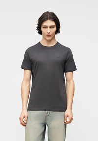 Νεαρός άνδρας με μεσαίου μήκους σκούρα μαλλιά, που φοράει απλό σκούρο γκρι κοντομάνικο μπλουζάκι και ανοιχτόχρωμο μπλε τζιν, στέκεται μπροστά σε ένα λευκό φόντο.