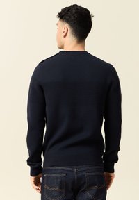 Maglione navy a maglia con texture a coste e design a righe sulla parte posteriore. Presenta maniche lunghe e collo rotondo. Indossato con jeans scuri.