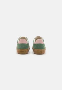 Zapatillas de ante verdes con acentos en rosa y suela marrón. El talón presenta un detalle en relieve con "1973". Diseño minimalista con una forma aerodinámica.