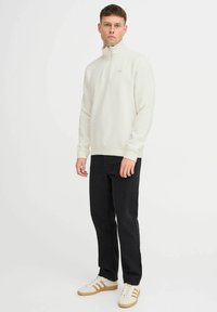 Sweatshirt en polaire crème clair à demi-zip avec des poignets côtelés, associé à un jean noir et des baskets beiges avec des rayures marron.