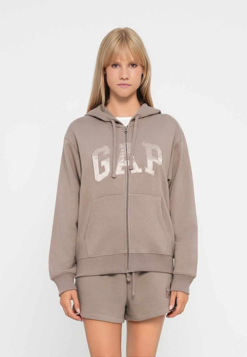 Brown zip-up hoodie featuring a large "GAP" logo in light beige. The fabric appears soft, with a relaxed fit and front pockets.