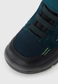 Dunkelblauer Schuh mit einer strukturierten Oberseite, ausgestattet mit einer schwarzen Gummisohle, elastischen Schnürsenkeln und einem grünen Akzentstich an der Seite.