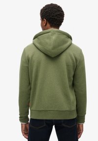 Felpa verde realizzata in tessuto morbido, dotata di cappuccio con cordino e polsini a costine. La parte posteriore presenta un design semplice con una forma leggermente aderente.