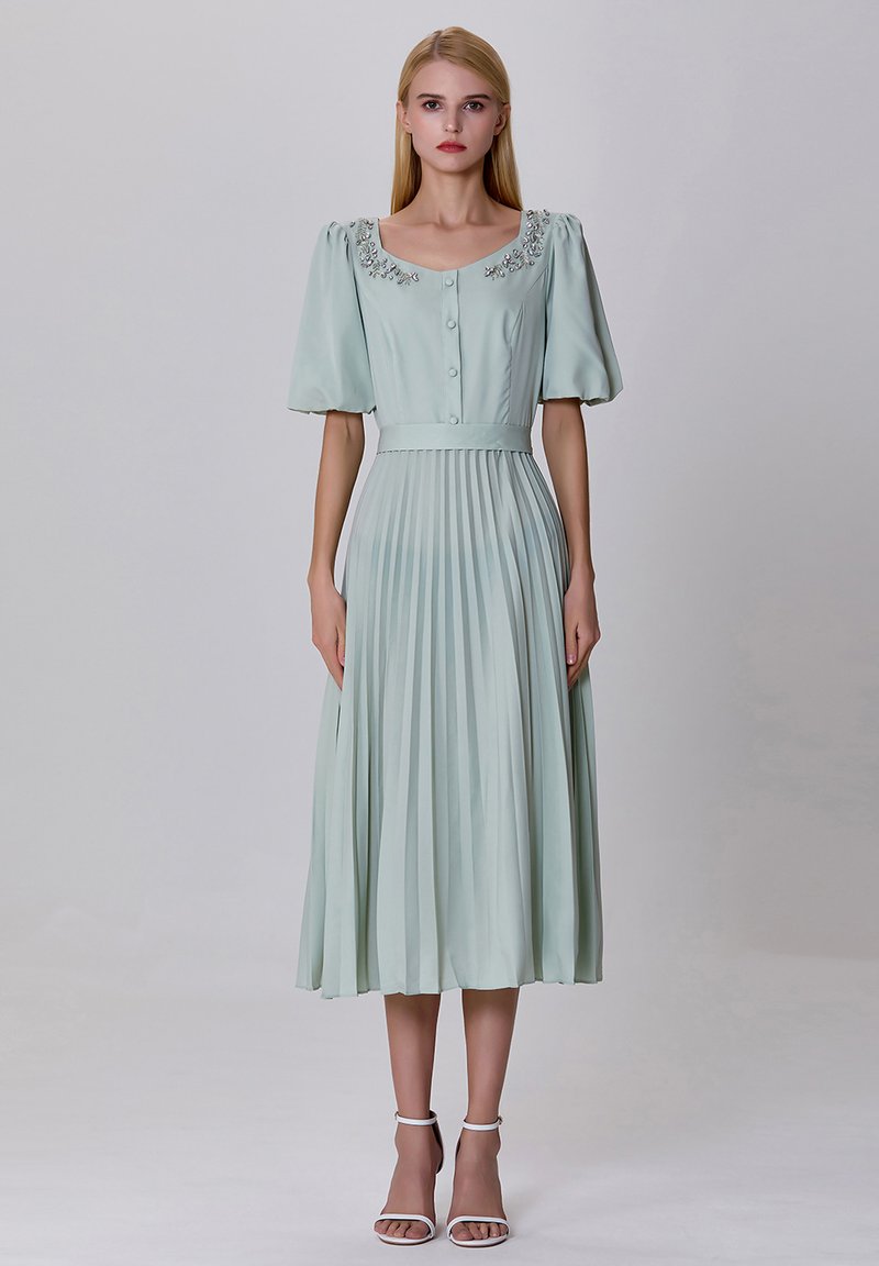 Robe vert menthe, longueur genou, avec manches bouffantes, devant boutonné et décolleté orné. Présente une jupe plissée et une ceinture à la taille ajustée.