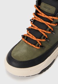 Sneakers Geox z zielonym zamszowym wierzchem, czarnymi akcentami, pomarańczowymi sznurówkami i gumową podeszwą. Posiadają metalowe oczka i teksturowane detale.