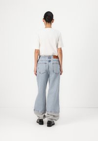 Šviesiai mėlynos plačios kelnės su šiek tiek išblukusiu efektu, su užpakalinėmis kišenėmis ir rudais odiniais ženklais ant juosmens, derinamos su baltu marškinėliu.