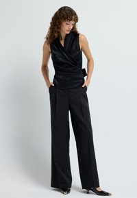 Top de satén negro con un cuello doblado, combinado con pantalones de pierna ancha negros y tacones negros puntiagudos con acentos dorados.